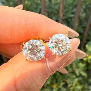 May include: A pair of gold stud earrings, each featuring a large, round, clear gemstone. The gemstones sparkle with multiple facets, reflecting light. The earrings are held up against a blurred background of green foliage and a brown railing.