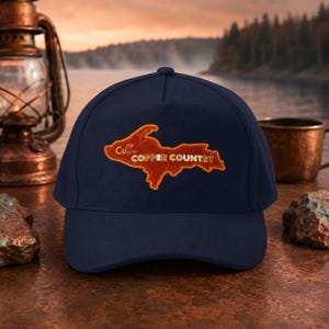 Puede incluir: Gorra de béisbol azul marino con un parche bordado que presenta el contorno naranja de una región y las palabras "Copper Country". El parche también incluye el símbolo químico del cobre, Cu.