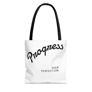 Może przedstawiać: Biała torba typu tote z czarnymi uchwytami. Torba zawiera napis "Progress" w kursywie, z napisem "Over Perfection" wydrukowanym poniżej mniejszą czcionką bezszeryfową. Torba wykonana jest z miękkiego materiału.
