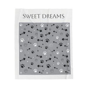 Op de afbeelding: Een witte deken met een grijs middenstuk met een patroon van zwarte en witte pootafdrukken en visgraten. De woorden "SWEET DREAMS" staan in het zwart bovenaan de deken.