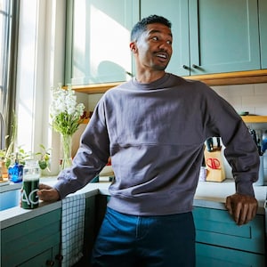 Pode incluir: Uma pessoa vestindo um moletom cinza e cal&ccedil;as azul escuro est&aacute; em uma cozinha. A pessoa est&aacute; segurando um copo com l&iacute;quido verde e as letras "AG" impressas. Flores brancas em um vaso na bancada.