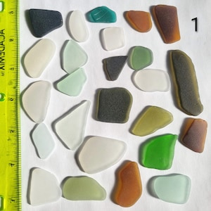 May include: A collection of twenty-six assorted pieces of smooth sea glass in various shapes and colors, including white, green, brown, and gray, arranged next to a yellow ruler showing measurements in inches, with the number 1 in the corner.