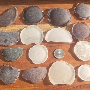 May include: A collection of fifteen smooth, weathered sea glass pieces in shades of dark brown, gray, and translucent white, arranged in a grid. A silver-colored United States quarter coin is placed among the pieces for scale reference.