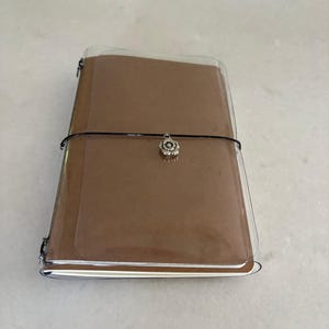 Puede incluir: Un cuaderno rectangular con cubierta marrón dentro de una funda protectora de plástico transparente. Una fina banda elástica negra rodea horizontalmente el centro del cuaderno, con un pequeño dije de flor de metal plateado sujeto a ella.