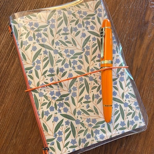 Peut inclure: Un carnet avec un motif floral de petites fleurs bleues et de feuilles vertes sur fond blanc, protégé par une couverture en plastique transparent. Un stylo-plume orange est fixé sur le devant du carnet par un élastique orange fin et horizontal.