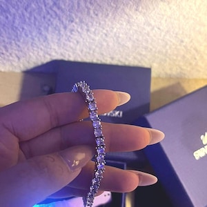 Può includere: Un braccialetto d'argento con numerose pietre preziose rotonde e trasparenti. Il braccialetto è tenuto contro uno sfondo sfocato, con un accenno di fonte di luce blu. Il gioiello è esposto vicino a una scatola blu scuro.