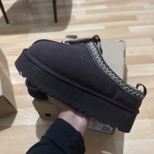 Puede incluir: Zapato tipo mocasín de ante marrón oscuro con suela gruesa negra. El zapato presenta un ribete decorativo cosido en blanco y beige, y la marca "UGG" está sutilmente grabada en el lateral. Talla 39.