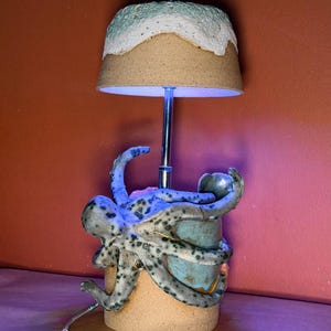 May include: A ceramic table lamp featuring a sculpted octopus with a speckled blue and gray finish wrapped around a cylindrical base. The lamp has a metallic central stem and a textured, dome-shaped shade with a light blue and white finish.