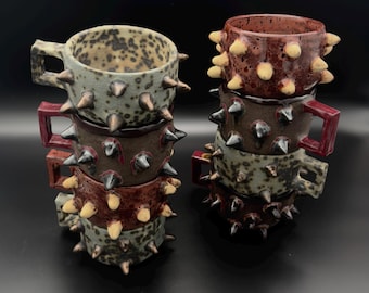 May include: Two stacks of ceramic mugs featuring protruding spikes on the exterior. The mugs come in speckled grey and solid reddish-brown glazes, each with a square-shaped handle. The spikes vary in color, appearing either light cream or dark.