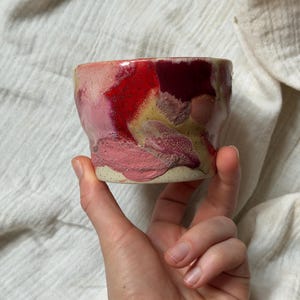 Può includere: Una piccola tazza in ceramica fatta a mano con un design astratto unico. La tazza presenta una miscela di colori, tra cui sfumature di rosa, rosso e giallo, creando un effetto marmorizzato. La base è di colore naturale non smaltato. Ideale per caffè o tè.