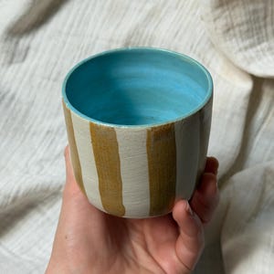 Op de afbeelding: Een handgemaakte keramische beker met een lichtblauwe binnenkant en verticale strepen in crème en bruin. De beker wordt in een hand gehouden tegen een witte stoffen achtergrond. De beker is cilindrisch en lijkt een drinkvat te zijn.
