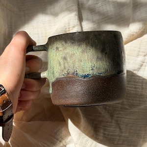 Könnte beinhalten: Eine handgefertigte Keramik-Tasse mit einzigartiger Glasur. Die obere Hälfte ist ein gesprenkeltes Grün, das in einen satten braunen Sockel übergeht. Die Tasse hat eine quadratische Form mit einem stabilen Henkel, ideal für Kaffee oder Tee.