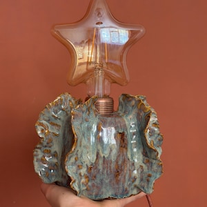 May include: A ceramic table lamp with a ruffled, wavy base featuring a mottled blue, brown, and tan glaze. The lamp includes a copper-colored socket and a clear, star-shaped LED filament light bulb that emits a warm glow from the center.