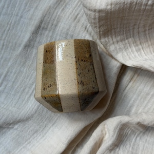 Puede incluir: Una taza de cerámica con un diseño geométrico. La taza presenta rayas verticales en tonos beige, marrón y beige claro. La taza tiene una textura moteada y una forma facetada. El fondo es una tela blanca texturizada.