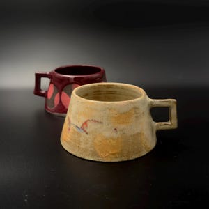 Puede incluir: Dos tazas de cerámica con cuerpos anchos y cónicos y asas cuadradas. La taza delantera es de color beige texturizado y moteado con manchas naranjas y marrones. La taza trasera es de color burdeos brillante con un patrón circular marrón oscuro.