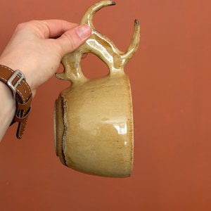 Puede incluir: Una taza de cerámica hecha a mano con un asa única en forma de cuerno. La taza tiene un esmalte de color canela con detalles marrones oscuros alrededor del borde y el asa. La taza se sostiene sobre un fondo naranja-marrón liso.