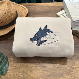 May include: A light beige sweatshirt featuring an embroidered design of a wolf's head with a red eye, held by a hand. The wolf is dark blue with black accents. The sweatshirt has Japanese text below the design.