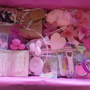 Puede incluir: Una caja de regalo llena de art&iacute;culos rosas que incluye un bolso beige acolchado, un peque&ntilde;o espejo con gr&aacute;ficos de tacones altos, esponjas de maquillaje, un objeto en forma de coraz&oacute;n, accesorios para el cabello, una cinta m&eacute;trica, un llavero y un corta&uacute;&ntilde;as.
