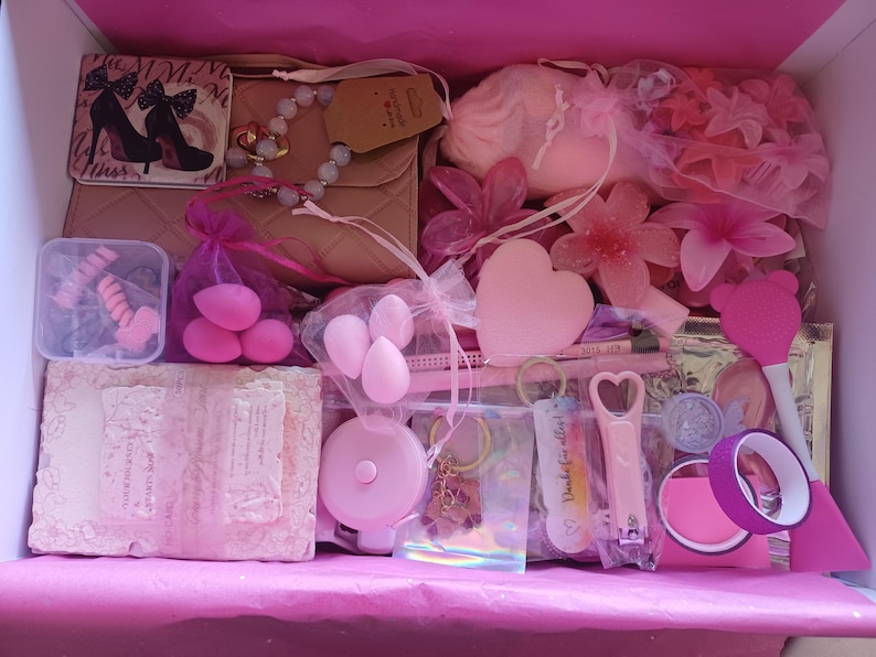 Puede incluir: Una caja de regalo llena de art&iacute;culos rosas que incluye un bolso beige acolchado, un peque&ntilde;o espejo con gr&aacute;ficos de tacones altos, esponjas de maquillaje, un objeto en forma de coraz&oacute;n, accesorios para el cabello, una cinta m&eacute;trica, un llavero y un corta&uacute;&ntilde;as.