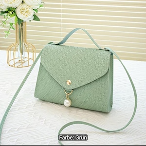 Puede incluir: Un bolso bandolera de color verde claro con una superficie texturizada, un asa superior y una correa larga para el hombro. El bolso cuenta con un cierre de solapa triangular asegurado con un bot&oacute;n dorado y un dije de perla colgante.