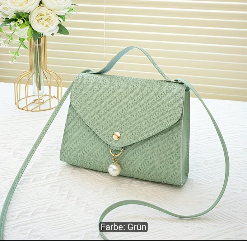Puede incluir: Un bolso bandolera de color verde claro con una superficie texturizada, un asa superior y una correa larga para el hombro. El bolso cuenta con un cierre de solapa triangular asegurado con un bot&oacute;n dorado y un dije de perla colgante.