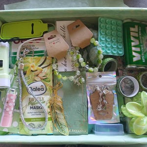 Puede incluir: Una caja de regalo llena de varios art&iacute;culos, incluyendo una linterna verde claro, una mascarilla facial de pistacho Balea, una pulsera de cuentas, un colgante de pluma dorado, una lata de mentas Vivil, un b&aacute;lsamo labial rosa con puntos y un adorno floral verde.