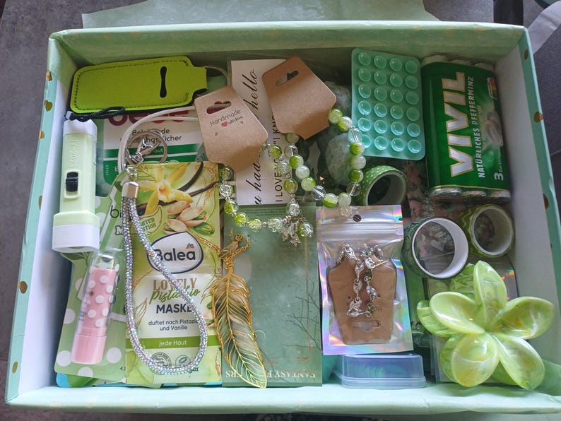 Puede incluir: Una caja de regalo llena de varios art&iacute;culos, incluyendo una linterna verde claro, una mascarilla facial de pistacho Balea, una pulsera de cuentas, un colgante de pluma dorado, una lata de mentas Vivil, un b&aacute;lsamo labial rosa con puntos y un adorno floral verde.