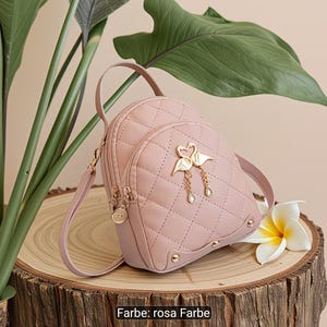 Puede incluir: Una mochila acolchada de color rosa claro con un emblema de metal dorado de dos cisnes que forman un coraz&oacute;n con dijes colgantes. La bolsa incluye un asa superior, correas ajustables y detalles de herrajes dorados en la base y cremalleras.
