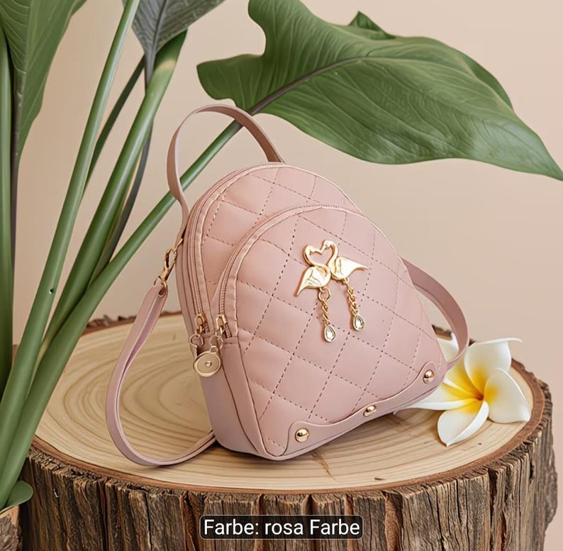 Puede incluir: Una mochila acolchada de color rosa claro con un emblema de metal dorado de dos cisnes que forman un coraz&oacute;n con dijes colgantes. La bolsa incluye un asa superior, correas ajustables y detalles de herrajes dorados en la base y cremalleras.