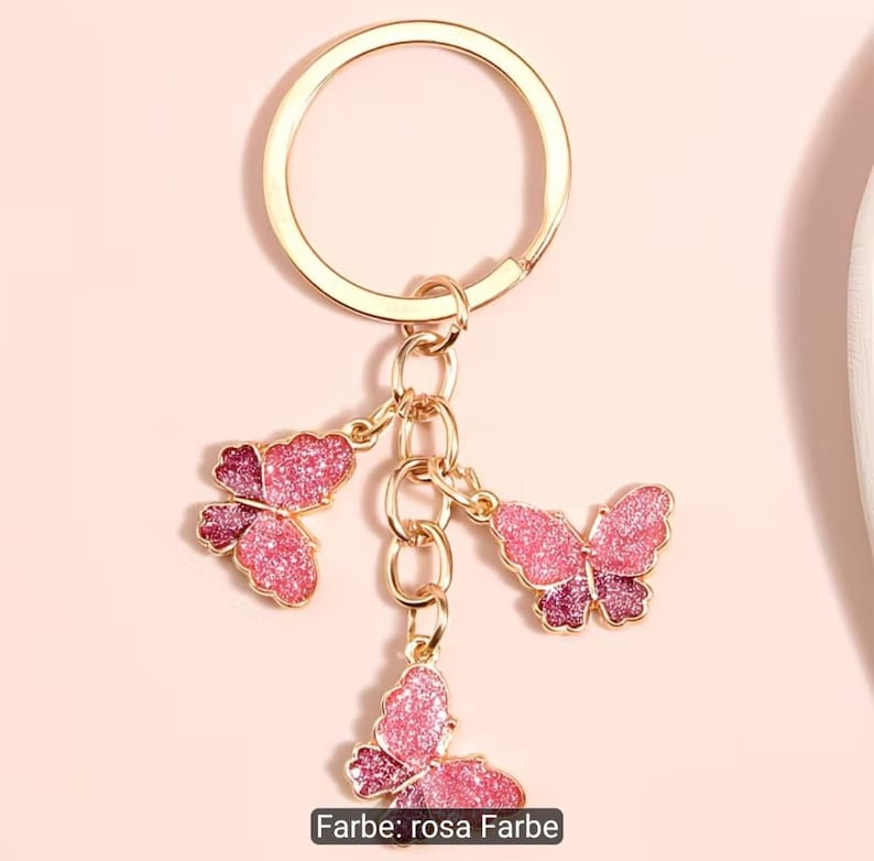 Puede incluir: Un llavero de metal dorado con tres dijes de mariposa colgantes. Cada mariposa est&aacute; decorada con un acabado degradado brillante en rosa y magenta. El llavero incluye una argolla circular unida a una cadena corta con las mariposas.