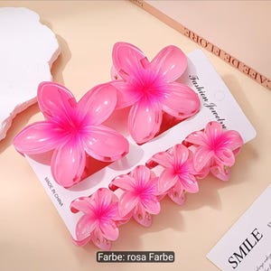 Puede incluir: Un conjunto de cinco pinzas para el cabello con forma de flor rosa sujetas a una tarjeta de presentaci&oacute;n blanca. Las pinzas presentan un dise&ntilde;o degradado con centros rosa oscuro y p&eacute;talos rosa claro. La tarjeta incluye el texto Fashion Jewelry.