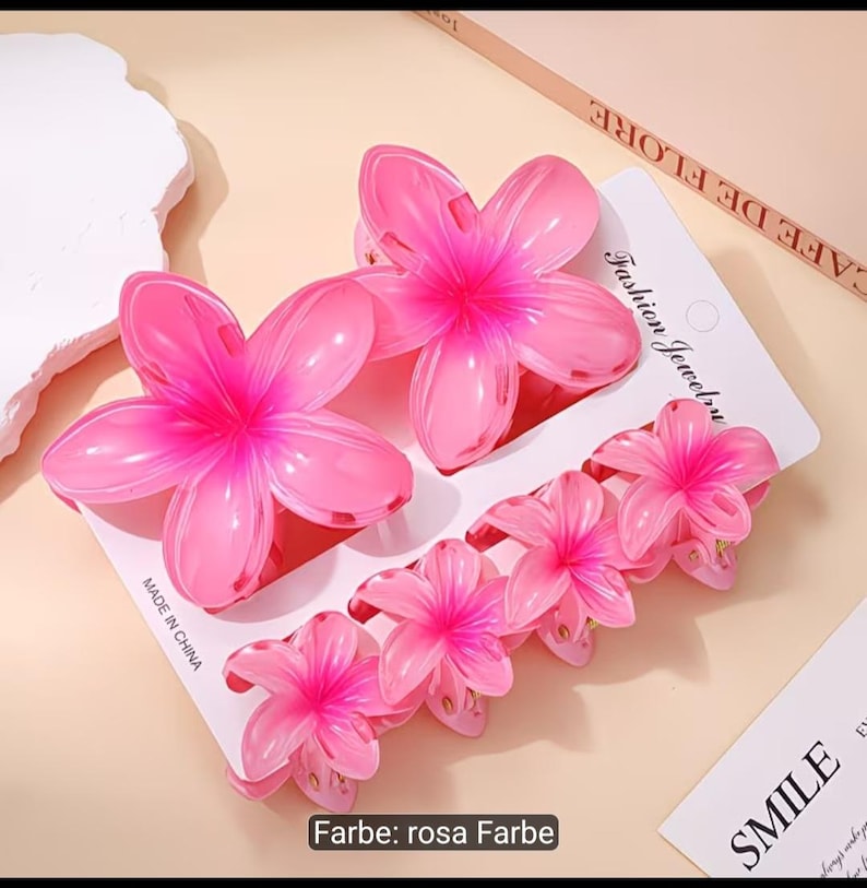Puede incluir: Un conjunto de cinco pinzas para el cabello con forma de flor rosa sujetas a una tarjeta de presentaci&oacute;n blanca. Las pinzas presentan un dise&ntilde;o degradado con centros rosa oscuro y p&eacute;talos rosa claro. La tarjeta incluye el texto Fashion Jewelry.