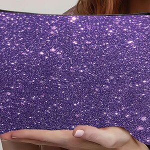 Puede incluir: Un estuche rectangular con cremallera que presenta un acabado de purpurina morada en toda la superficie. El exterior muestra una textura densa y brillante con numerosas part&iacute;culas que reflejan la luz, creando un efecto centelleante. Un tirador de cremallera de metal plateado est&aacute; sujeto en la esquina superior.