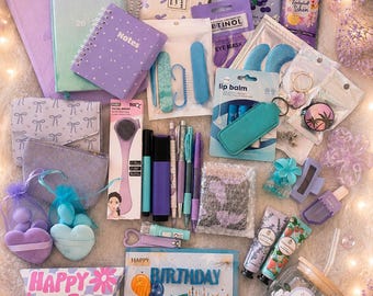 Puede incluir: Una colección de artículos de papelería y belleza que incluye un cuaderno de espiral morado, un cepillo facial, bálsamo labial, limas de uñas, cremas de manos, un vaso de cristal con tapa y una tarjeta con el texto Happy Birthday to You.