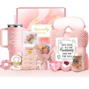 Puede incluir: Un set de regalo rosa que incluye un vaso con texto en alem&aacute;n, una manta de felpa con la etiqueta DIESE DECKE ALS EINE Umarmung VON MIR F&Uuml;R DICH, un antifaz, un ramo de flores de ganchillo, una vela, un coletero y un frasco de sales de ba&ntilde;o.