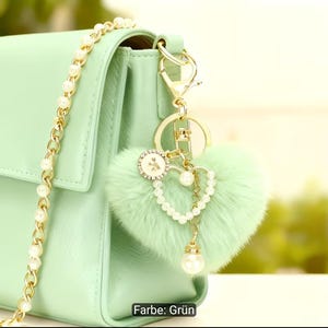 Puede incluir: Un bolso de mano verde claro con una correa de cadena dorada con perlas blancas. Un llavero de piel sint&eacute;tica en forma de coraz&oacute;n verde claro cuelga del lateral, decorado con un anillo dorado, un peque&ntilde;o dije circular y una perla blanca.