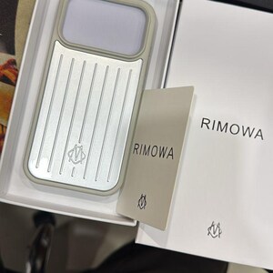Può includere: Una custodia per telefono color argento e beige con un motivo a scanalature verticali e un piccolo logo, presentata in una scatola bianca insieme a una carta beige abbinata e un libretto bianco, entrambi con il testo RIMOWA e un piccolo logo nero.