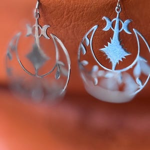 May include: A pair of silver-colored earrings. Each earring features a crescent moon design with a central starburst and floral accents. The earrings hang from a simple hook, set against an orange background.
