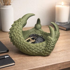 May include: A green, claw-shaped trinket dish, holding keys. The dish is textured with scale-like details and has three prominent claws. The dish sits on a wooden surface, with a candle and vase in the background.