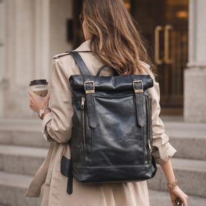 May include: A black roll-top backpack featuring a textured finish, two vertical front pockets with brass-toned zippers, and two adjustable straps with brass-toned buckles. The bag includes a top handle and side zippers for access to the main compartment.
