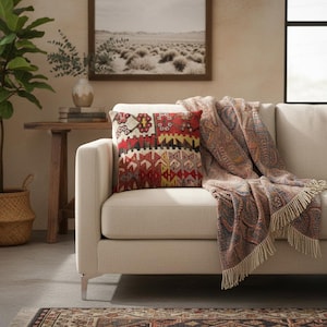 Peut inclure: Un canapé crème avec une couverture à motifs et un coussin décoratif. Le coussin présente un motif géométrique rouge, jaune et crème. Une photo encadrée de paysage est accrochée au-dessus du canapé.