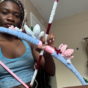 May include: A handcrafted bow and arrow set constructed from pipe cleaners in shades of light blue, white, pink, and dark red. The bow features decorative pipe cleaner flowers in white and pink, while the arrow has a dark red tassel at the end.