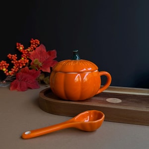 Może przedstawiać: Jasnopomarańczowy ceramiczny kubek w kształcie dyni z pasującą pokrywką z ciemnozieloną łodygą i wytłoczonym słowem PUMPKIN na górze. Mała pomarańczowa ceramiczna łyżeczka w kształcie dyni spoczywa przed kubkiem na drewnianej tacy.