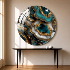 May include: A large, round wall art piece with a glossy finish. The artwork features a teal, gold, and white agate design. The piece is mounted on a beige wall above a black table with three legs. Two small, white decorative spheres sit on the table.