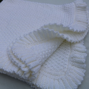Könnte beinhalten: Eine weiße gehäkelte Babydecke mit einem gekräuselten Rand.