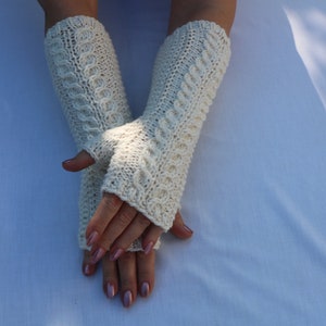 May include: A pair of white knitted fingerless gloves with a cable knit pattern. The gloves are folded over the hands and the fingertips are visible.