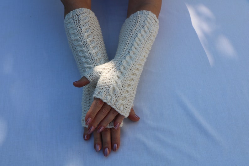 May include: A pair of white knitted fingerless gloves with a cable knit pattern. The gloves are folded over the hands and the fingertips are visible.