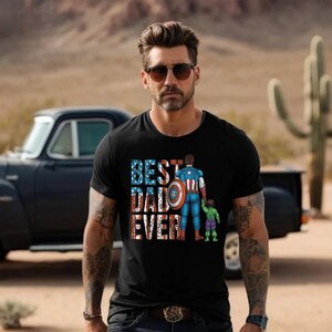 May include: Black t-shirt with the text "BEST DAD EVER" in red, white, and blue. The graphic depicts a superhero dad holding hands with a child dressed as the Hulk. The background shows a desert landscape.