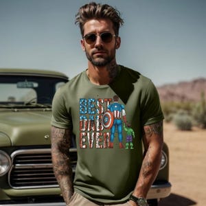 May include: Olive green t-shirt with the text "BEST DAD EVER" in red, white, and blue. The graphic features a superhero dad and two children. The shirt is short-sleeved.