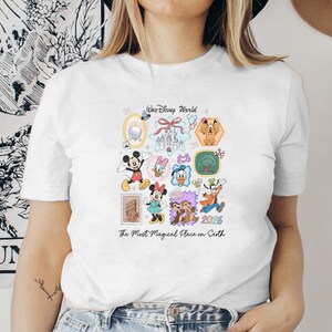 Peut inclure: T-shirt blanc orn&eacute; d'illustrations color&eacute;es de personnages et de lieux de Disney, dont Mickey Mouse, Minnie Mouse et le ch&acirc;teau de Cendrillon. Le texte "Walt Disney World" et "The Most Magical Place on Earth" sont &eacute;galement pr&eacute;sents.